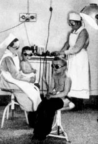 Ein Kind das auf einem Hocker sitzt. Dahinter sitzt eine Krankenschwester mit einem Kind auf dem Schoß und eine weitere Krankenschwester steht dahinter an einem Tisch